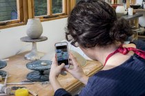 Blick über die Schulter einer Frau, die in der Keramikwerkstatt sitzt und ihr Handy checkt. — Stockfoto