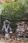 Homem barbudo de gorro preto e parka em pé no jardim no outono, usando machado para cortar pedaço de madeira no bloco de corte . — Fotografia de Stock