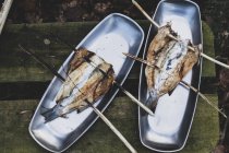 Высокоугольный крупный план жареной рыбы на деревянных шампурах на металлических пластинах на открытом воздухе — стоковое фото