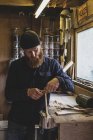 Hombre barbudo con gorro negro de pie en el banco de trabajo en el taller, trabajando en pieza de madera . - foto de stock