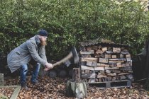 Homme barbu en bonnet noir et parka debout dans le jardin en automne, en utilisant la hache pour couper un morceau de bois sur le bloc de coupe
. — Photo de stock