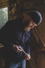 Bearded man wearing black beanie standing in workshop, examining piece of wood. — Stock Photo