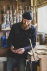 Bearded man wearing black beanie standing next to workbench in workshop, checking mobile phone. — Stock Photo