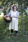Woman in apron holding brown wicker basket with freshly picked apples, smiling in camera. — Stock Photo