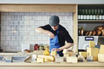 Cheesemonger cutting cheese with cheese wire surrounded by variety of cheese on counter top — Stock Photo