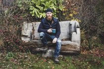 Bearded uomo in berretto nero seduto su una panchina di legno in giardino, tenendo tazza blu, guardando in macchina fotografica . — Foto stock