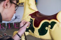 Close-up of woman in glasses in workshop painting traditional wooden horse from merry-go-round. — Stock Photo