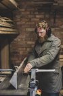 Bearded man standing in workshop, wearing ear protectors, working on piece of wood. — Stock Photo