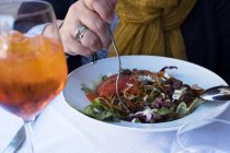 Gros plan d'une femme assise à une table de café et tenant une fourchette dans une salade mixte et un verre de vin frais . — Photo de stock