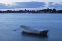 Размытие движения, когда лодка швартуется у берега в спокойной воде с горизонтом города на расстоянии с высокими зданиями и огнями, Коппель, Дания — стоковое фото