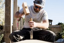 Hombre con gorra de béisbol y gafas de sol en el sitio de construcción, usando mazo y cincel, trabajando en viga de madera . - foto de stock