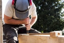 Hombre con gorra de béisbol y protectores de oídos en el sitio de construcción, trabajando en viga de madera . - foto de stock