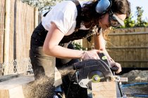 Woman wearing protective goggles and ear protectors holding circular saw, cutting piece of wood on building side. — Stock Photo
