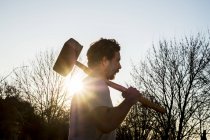 Bärtiger Mann, der bei Sonnenuntergang draußen spaziert und einen großen Holzhammer im Gegenlicht trägt. — Stockfoto