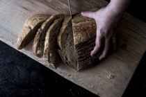 Person hand holding loaf of bread and using knife to cutting slices. — Stock Photo