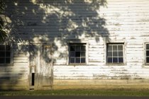 Granja antigua con revestimiento de madera pelada y ventanas con sombra de árbol . - foto de stock