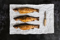 High angle close-up of three fresh smoked whole trouts and knife on white paper. — Stock Photo