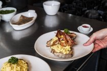 High angle close-up of person holding plate with scrambled eggs and crispy bacon on slice of bread. — Stock Photo
