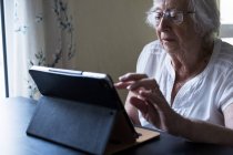 Старша жінка сидить за столом і використовує цифровий планшет з сенсорним екраном . — стокове фото