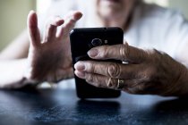 Close-up de mãos de mulher idosa sentada à mesa e usando telefone celular . — Fotografia de Stock