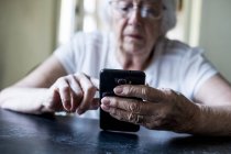 Primo piano delle mani della donna anziana seduta a tavola e che utilizza il telefono cellulare . — Foto stock
