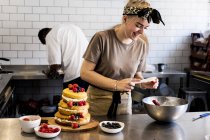 Cocinera femenina trabajando en cocina comercial montando torta de esponja en capas con frutas frescas . - foto de stock
