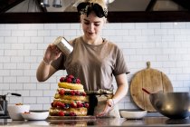 Female cook working in commercial kitchen sprinkling icing sugar over layered cake with fresh fruits. — Stock Photo