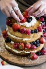 Hände einer Köchin, die Kuchenschichten mit frischer Sahne und frischen Früchten, Erdbeeren und Blaubeeren zusammenfügt. — Stockfoto
