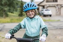 Retrato de menino de idade elementar vestindo casaco de chuva e capacete straddling bicicleta . — Fotografia de Stock