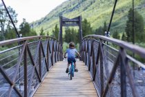 Visão traseira do menino idade elementar na montanha atravessando ponte estreita . — Fotografia de Stock