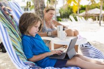 Preschooler boy using laptop with mother at beach. — Stock Photo