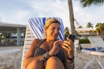 Donna matura che utilizza smartphone sulla spiaggia al tramonto, Grand Cayman Island — Foto stock