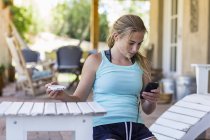 Ragazza adolescente bionda utilizzando smartphone mentre dipinge mobili da esterno in bianco . — Foto stock