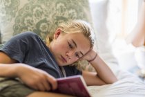Adolescente chica acostada en la cama y leyendo por la luz de la ventana . - foto de stock