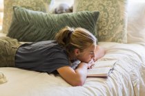 Adolescente chica acostada en la cama y leyendo por la luz de la ventana . - foto de stock