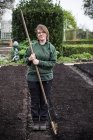 Smiling woman holding wooden rake standing on freshly laid bed of soil in vegetable garden. — Stock Photo