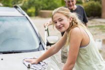 Teenage granddaughter looking in camera as washing car together with senior grandfather in driveway — Stock Photo