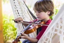 Маленький хлопчик грає на скрипці зовні в саду на гамаку — стокове фото
