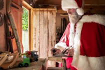 Mann im Weihnachtsmann-Kostüm steht in Werkstatt und baut Spielzeugauto aus Holz. — Stockfoto