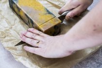 High angle close-up of female hands cutting yellow and black homemade bar of soap with kitchen knife. — Stock Photo