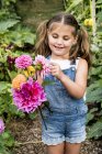 Mädchen in Jeans-Latzhose steht in einem Garten und hält rosa Dahlien in der Hand. — Stockfoto