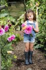 Mädchen in Jeans-Latzhose steht im Garten und hält rosa Dahlien in der Hand. — Stockfoto