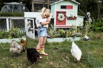 Chica rubia de pie en el jardín frente al gallinero, sosteniendo pollo blanco . - foto de stock
