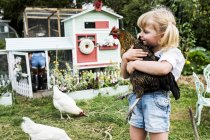 Blondes Mädchen steht im Garten vor Hühnerstall und hält braunes Huhn. — Stockfoto