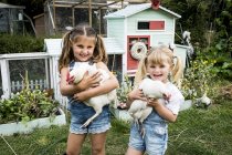 Two girls standing in front of hen house in garden, holding white chickens, smiling in camera. — Stock Photo