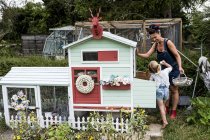 Sorrindo mulher e menina de pé no jardim ao lado de galinha colorida casa . — Fotografia de Stock