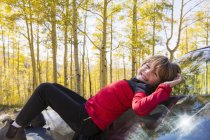 Lächelnder Junge im Grundschulalter liegt auf Motorhaube eines blauen Geländewagens im Wald. — Stockfoto