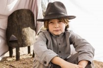 Хлопчик початкового віку в капелюсі сидить у наметі на відкритому повітрі з листів — стокове фото