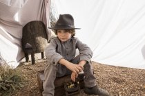 Мальчик младшего возраста в шляпе сидит на чемодане в наружной палатке из простыней — стоковое фото