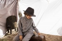 Ragazzo in età elementare che indossa cappello giocando con valigia in tenda esterna fatta di lenzuola — Foto stock